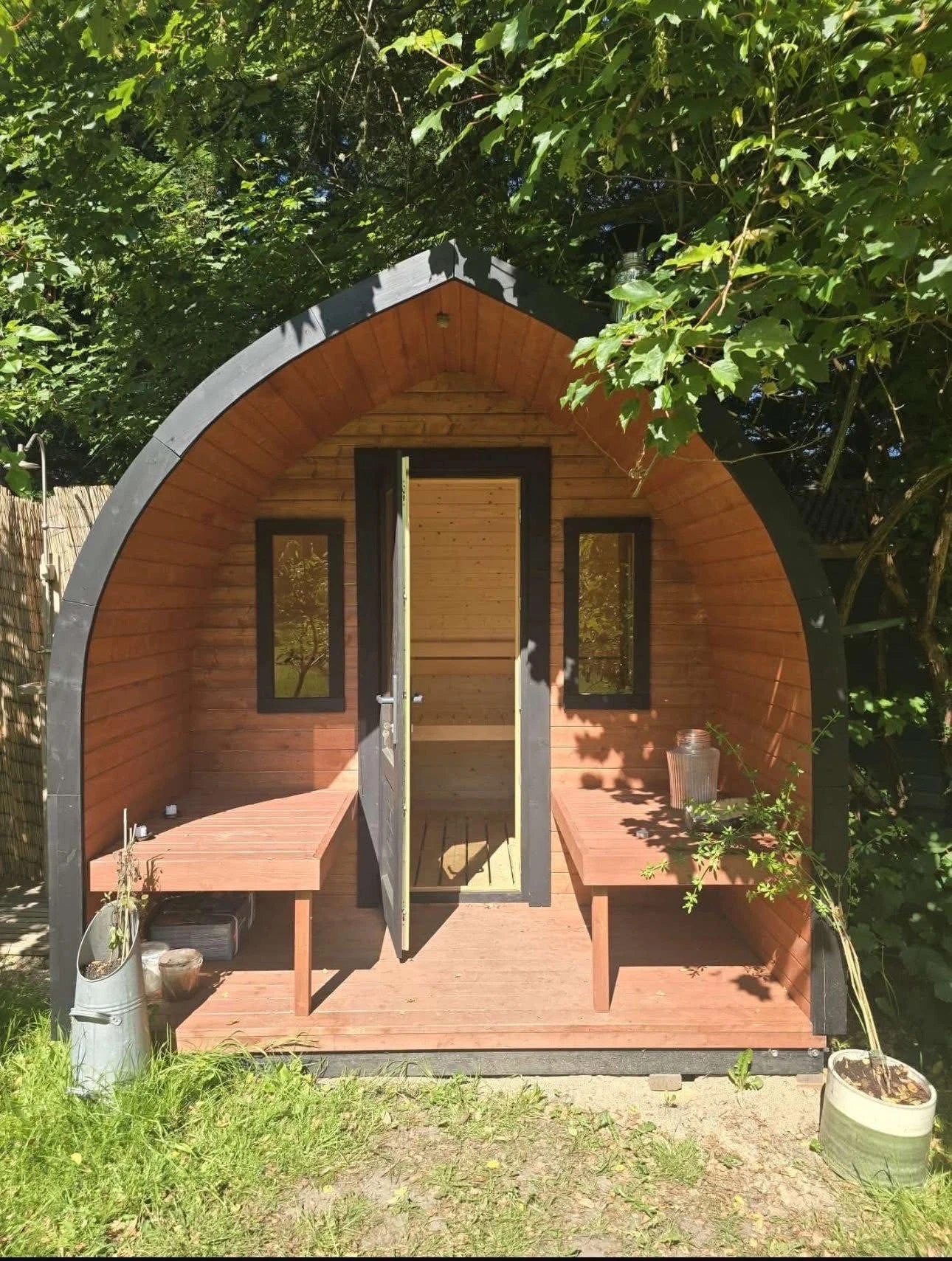 Small wooden glamping pod with a curved roof, two narrow windows, and a central door opening onto a small porch with built-in benches on each side, surrounded by green trees and sunlight filtering through the leaves.