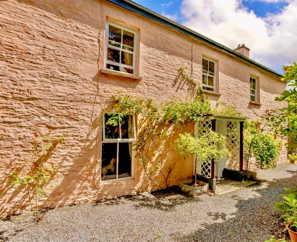 Traditional two-storey cottage with light pink stone walls, white-framed sash windows, and a small porch covered by a trellis and climbing plants, surrounded by greenery and a gravel path under a bright blue sky.